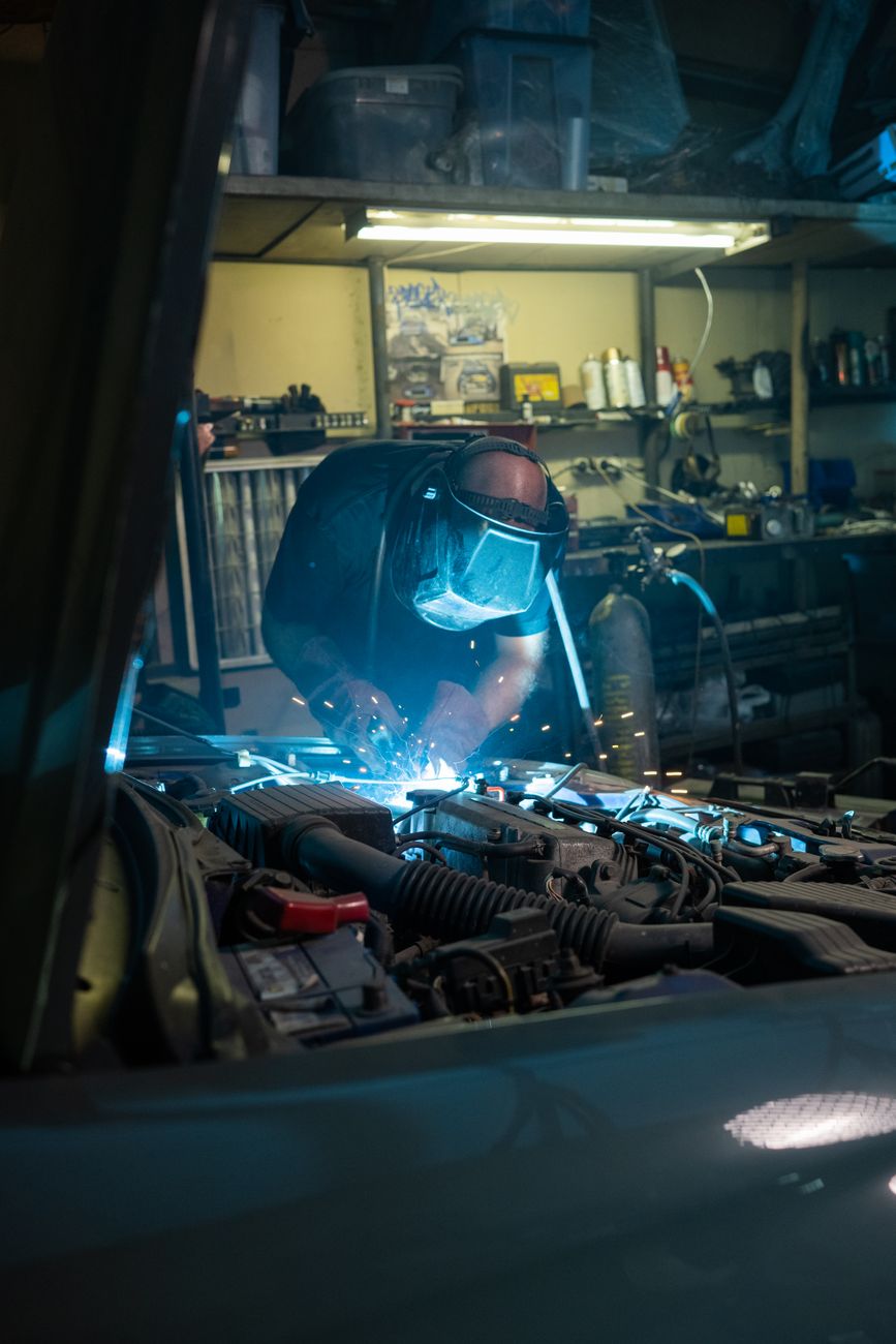 Mechanic wearing safety gear welding under hood of a car in a garage workshop.